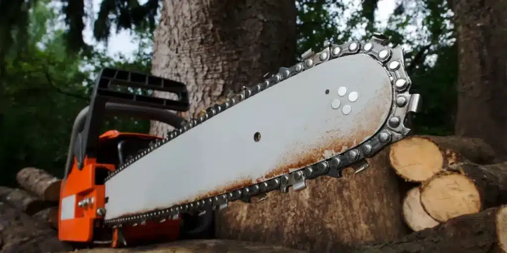 A close-up view of a chainsaw placed on a pile of logs. The focus is on the rusty chain and blade, while the orange body of the chainsaw is partially visible. Large tree trunks in the background suggest recent land clearing or stump grinding activities.